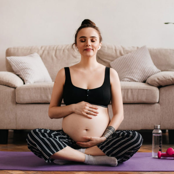cheerful-brunette-pregnant-woman-enjoys-music-white-wireless-headphones-gently-touches-belly-cheerful-tanned-girl-black-pants-top-meditating-purple-carpet